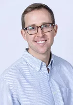 Headshot of a smiling person with short light brown hair and glasses, wearing a light blue striped button-up shirt against a light gray background.