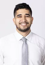 Smiling person with short curly dark hair and trimmed facial hair, wearing a white dress shirt and a striped gray tie against a light gray background.