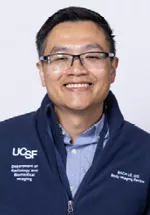 Headshot of a smiling person with short black hair and glasses, wearing a navy UCSF zip-up jacket over a light blue collared shirt against a light gray background.