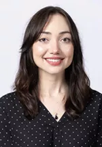 Smiling person with shoulder-length dark brown hair, wearing a black blouse with a subtle dot pattern against a light gray background.