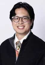 Headshot of a smiling person with medium-length dark hair and round glasses, wearing a white shirt, patterned tie, and black zip-up sweater against a light gray background.