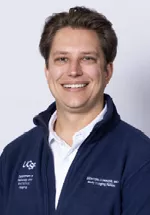 Headshot of a smiling person with short light brown hair, wearing a navy UCSF zip-up jacket over a light shirt against a light gray background.