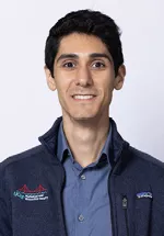 Smiling person with short dark hair, wearing a navy vest over a blue button-up shirt against a light gray background.