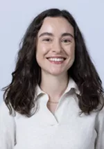 Smiling person with long dark wavy hair, wearing a white collared blouse against a light gray background.