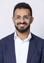 Smiling person with short curly hair, a beard, and glasses, wearing a navy blazer over a white shirt against a light gray background.