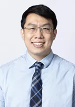 Smiling person wearing glasses, a light blue dress shirt, and a blue plaid tie against a light gray background.