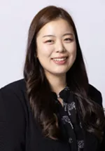 Smiling person with long dark hair, wearing a black blazer over a patterned blouse against a light gray background.