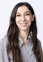 Smiling person with long dark hair, wearing a striped button-up shirt against a light gray background.
