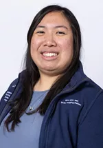 Headshot of a smiling person with long straight dark hair, wearing a navy UCSF zip-up jacket against a light gray background.
