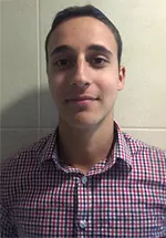 Head-and-shoulders photo of a person wearing a red, blue, and white checkered button-down shirt, facing the camera against a light tiled wall.