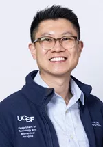Headshot of a smiling person with short dark hair and glasses, wearing a light blue collared shirt under a navy UCSF zip-up jacket against a light gray background.