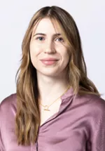Person with long blonde hair, wearing a mauve satin blouse and a necklace against a light gray background.