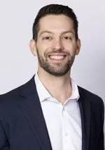 Smiling person with short dark hair and trimmed facial hair, wearing a navy blazer over a white shirt against a light gray background.
