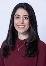 Smiling person with long dark hair, wearing a burgundy sweater and a necklace against a light gray background.
