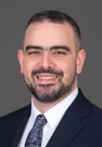 Professional headshot of a smiling man with short dark hair and a neatly trimmed beard, wearing a dark navy suit, white dress shirt, and patterned tie against a plain gray background.