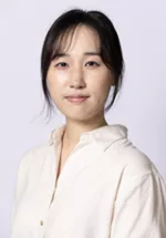 Person with dark hair pulled back, wearing a cream-colored blouse against a light gray background.