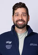 Professional headshot of a smiling person with short dark hair and a beard, wearing a navy UCSF zip-up jacket over a light gray shirt against a light gray background.
