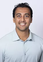 Smiling person with short curly dark hair, wearing a light blue polo shirt against a light gray background.