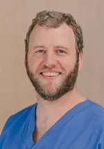 Headshot of a smiling man with short brown-and-gray hair and a beard, wearing blue medical scrubs against a neutral beige background.