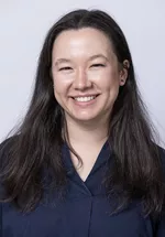 Smiling person with long dark hair, wearing a navy blouse against a light gray background.