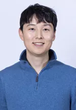 Smiling person with short dark hair, wearing a blue quarter-zip pullover against a light gray background.