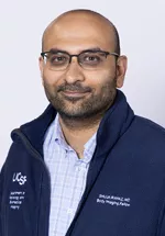 Headshot of a smiling bald person with glasses and closely trimmed facial hair, wearing a light blue striped shirt under a navy UCSF zip-up jacket against a light gray background.