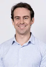 Smiling person with short brown hair, wearing a light blue checkered button-up shirt against a light gray background.
