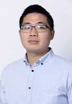 Person wearing glasses and a light blue button-up shirt against a light gray background.