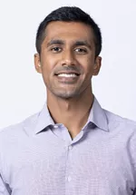 Smiling person with short dark hair, wearing a light lavender button-up shirt against a light gray background.