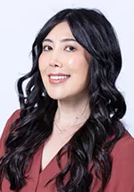 Smiling person with long dark wavy hair, wearing a burgundy blouse against a light gray background.