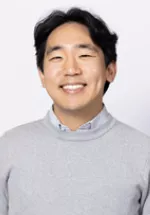 Smiling person with medium-length dark hair, wearing a light gray sweater against a light gray background.