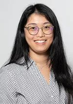 Headshot of a smiling person with long straight dark hair and round glasses, wearing a light gray patterned blouse and a delicate necklace against a light gray background.