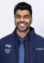Headshot of a smiling person with short dark hair and a beard, wearing a navy UCSF zip-up jacket, dark patterned shirt, and blue tie against a light gray background.