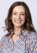 Smiling person with shoulder-length wavy brown hair, wearing a patterned blouse against a light gray background.