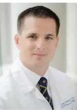 Head-and-shoulders portrait of a man wearing a white medical coat, white shirt, and patterned tie, facing the camera against a softly blurred light background.