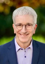 Head-and-shoulders portrait of an older man with short gray hair and round glasses, smiling at the camera. He wears a navy blazer over a light lavender shirt, with softly blurred green and golden foliage in the background.