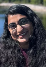Portrait of Sangati Pancholi wearing glasses and smiling outdoors, with long dark hair and greenery in the background.