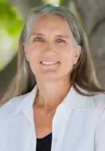 Headshot of a smiling woman with long gray hair, wearing a white collared shirt over a black top, photographed outdoors against a softly blurred green background.