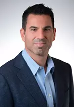 Professional headshot of a man with short dark hair and a trimmed goatee, wearing a blue blazer over a light blue dress shirt. He is facing the camera with a slight smile against a light gray studio background.