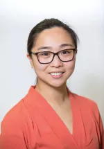 Professional headshot of a woman with long dark hair and round glasses, wearing a patterned blouse and smiling at the camera against a light background.