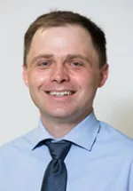 Professional headshot of a smiling man wearing a light blue dress shirt and a blue tie, facing the camera. The portrait is set against a neutral light gray background and has a polished appearance suitable for a faculty, physician, or professional profile.