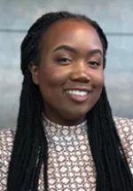 Professional headshot of a smiling woman with long braided hair, facing the camera. She is wearing a patterned blouse and is photographed against a neutral gray background, creating a polished portrait suitable for a professional profile.