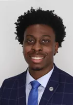 Professional headshot of a smiling man with short curly hair, facing the camera. He is wearing a blue checkered suit jacket, white dress shirt, and blue tie. The portrait is set against a neutral background and has a polished professional appearance.
