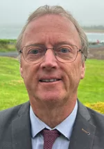 Head-and-shoulders portrait of a person wearing glasses, a gray suit jacket, light blue dress shirt, and burgundy tie, smiling outdoors with a grassy area and waterfront in the background.