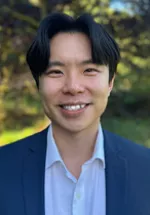 Professional headshot of a smiling man with dark hair, facing the camera. He is wearing a navy blazer over a white dress shirt. The portrait is taken outdoors with a softly blurred background of greenery and natural light.