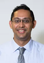 Professional headshot of a smiling man wearing glasses, a light dress shirt, and a patterned tie. He is facing the camera against a light gray background, creating a polished and professional portrait suitable for a faculty or physician profile.