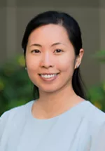 Professional headshot of a smiling woman with dark hair pulled back, facing the camera. She is wearing a light blue blouse, and the background is softly blurred with greenery, creating a warm and approachable professional portrait.