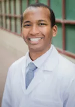Headshot of a man in a white medical coat smiling, standing outdoors with a blurred building background.