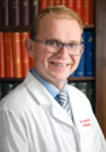 Headshot of a man wearing glasses and a white medical coat, smiling in front of a bookshelf.