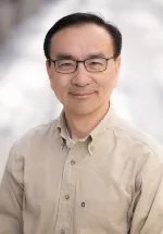 Professional headshot of a man wearing glasses and a light tan button-down shirt. He is smiling gently at the camera against a softly blurred gray and white background, creating a warm and approachable portrait.